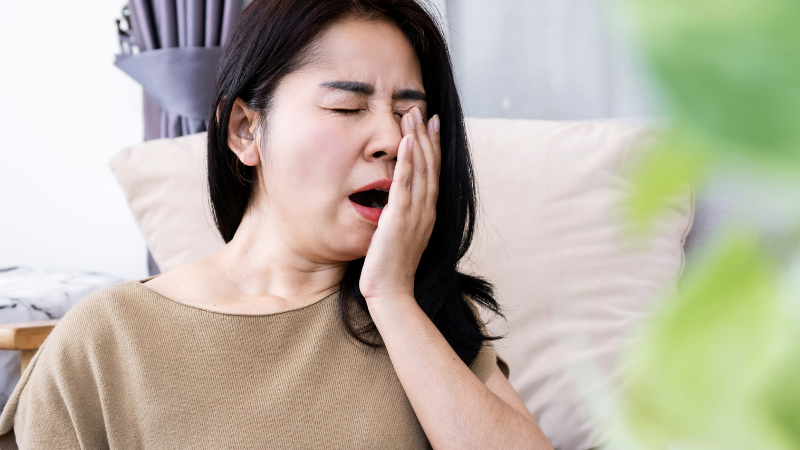 A woman sitting on a beige couch, yawning with her hand covering her mouth, conveying tiredness. Soft lighting and a blurred green plant in the foreground.