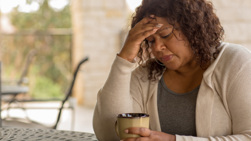 A woman sits at a table, looking pensive and weary, with her hand on her forehead. She holds a coffee mug, conveying a sense of contemplation and fatigue.